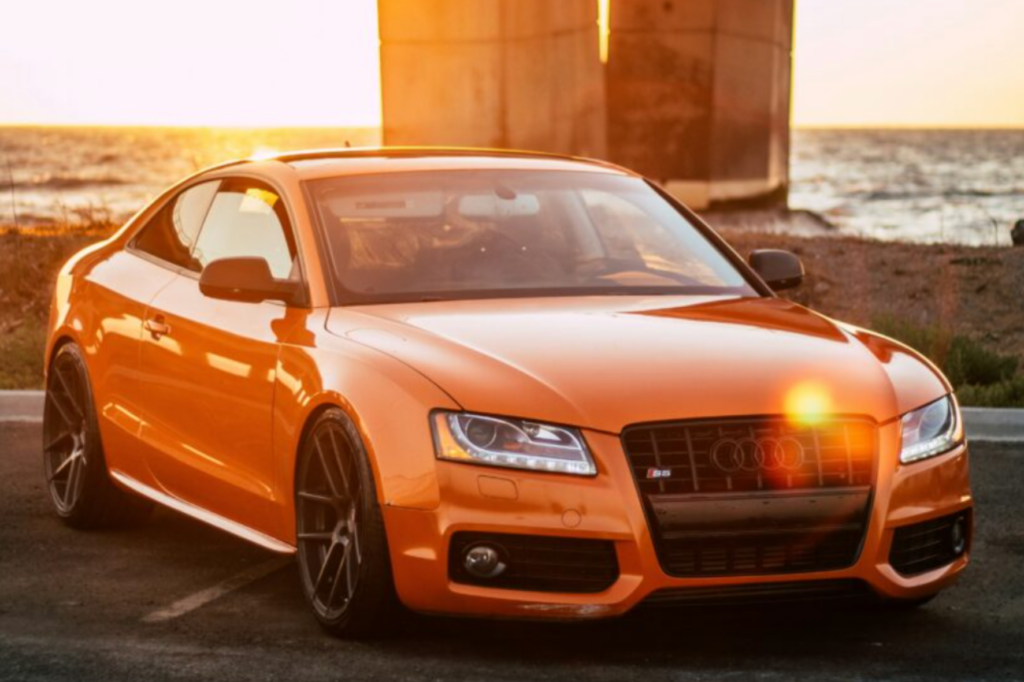 Orange Audi car parked in front of ocean, waves crashing in background.