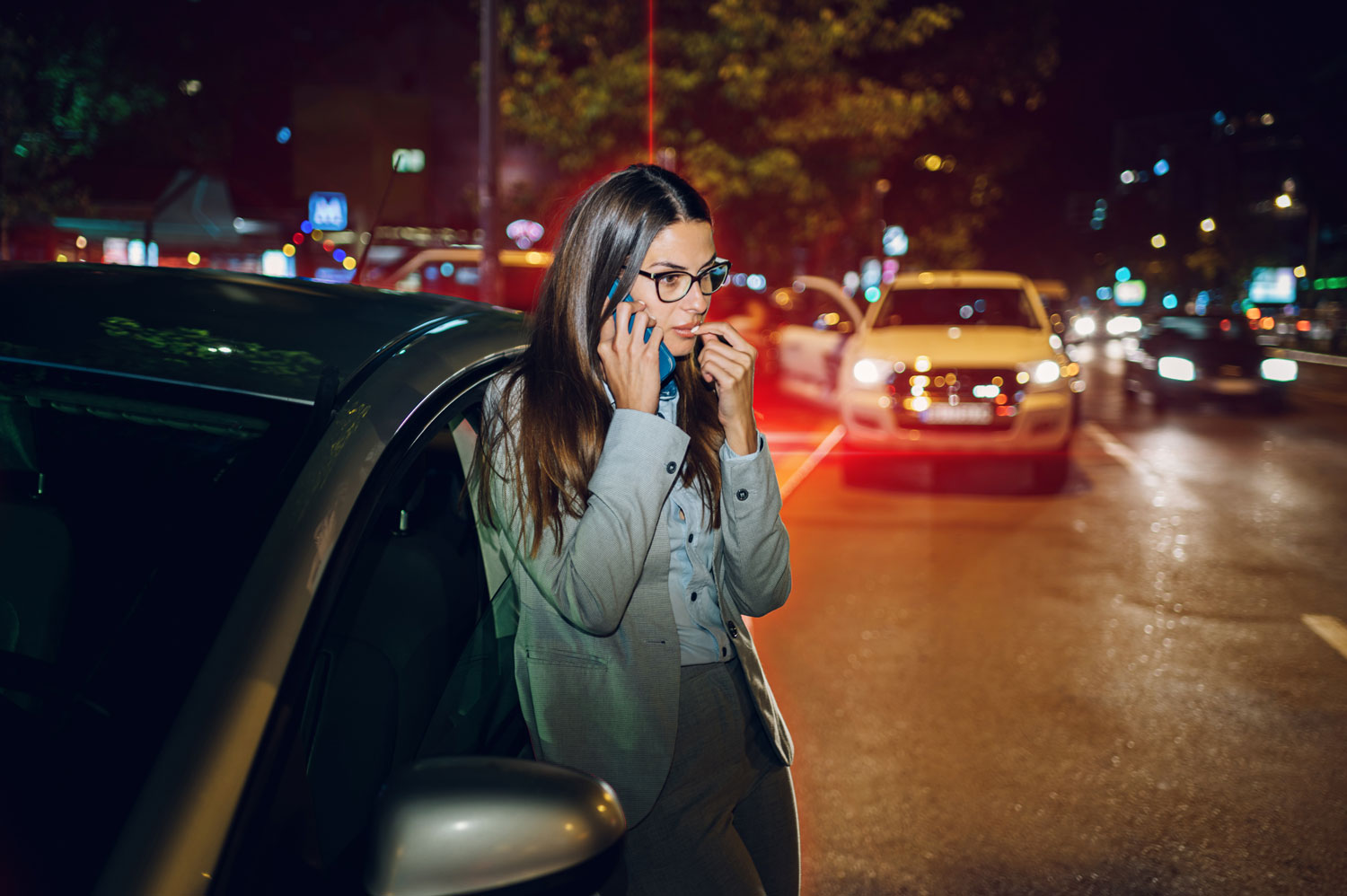 A woman next to her car with hazard lights on, pulled over on a highway at night, signaling for roadside assistance.
