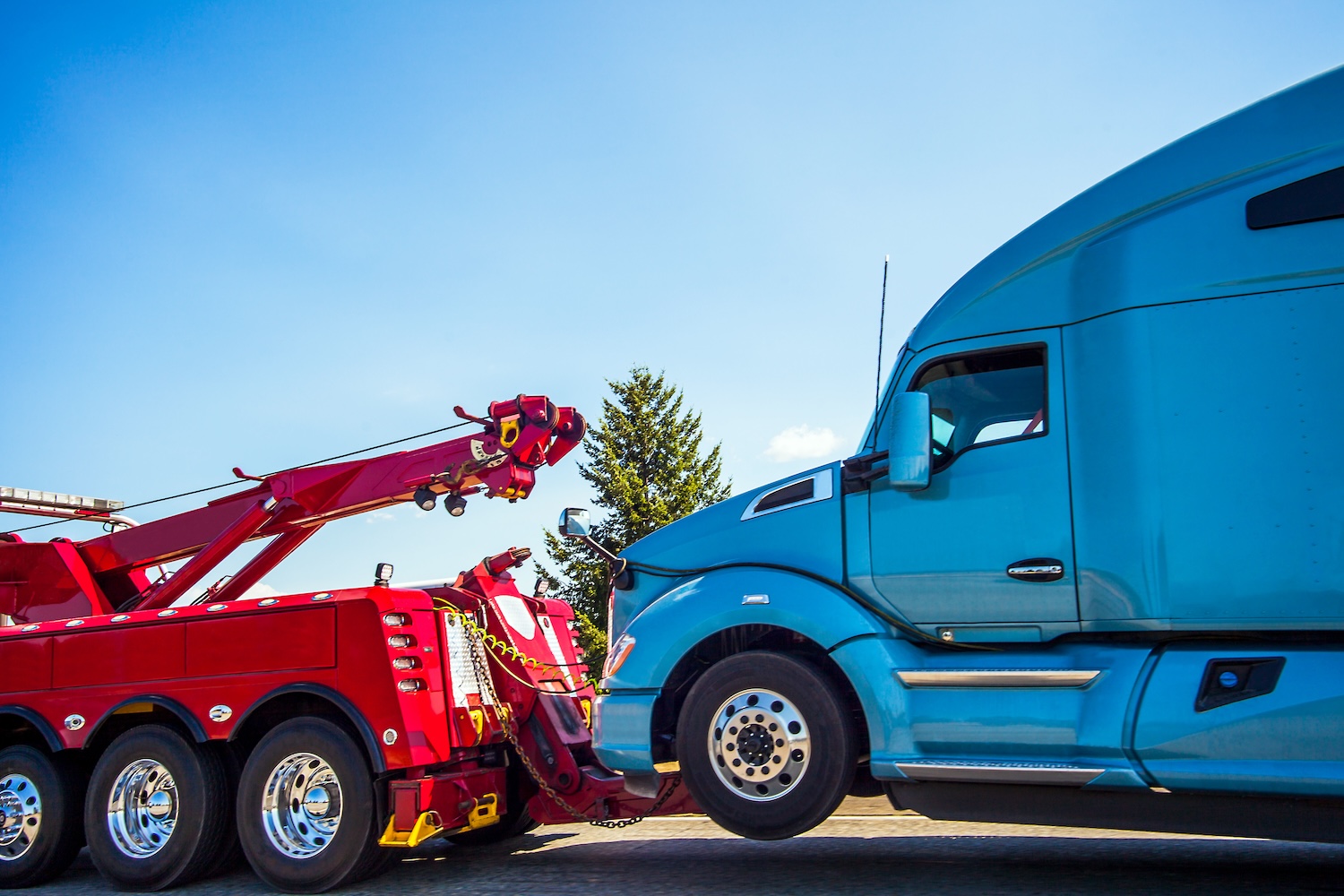  Heavy duty tow truck lifting a large blue semi-truck on the side of the road under a clear blue sky.