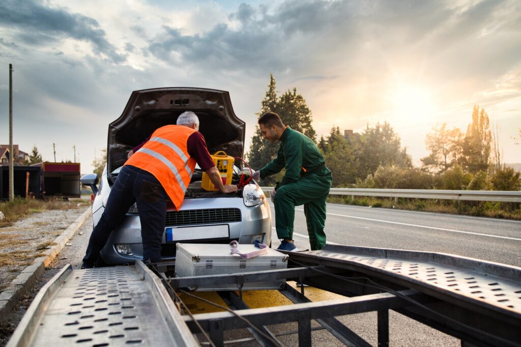 Professional towing technicians providing roadside assistance and jump-starting a vehicle safely on the side of the road at sunset.
